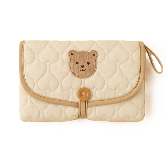 Tapis à langer plié en coton matelassé beige avec motif ourson brodé, fermeture bouton bois et liseré beige, posé sur fond blanc.
